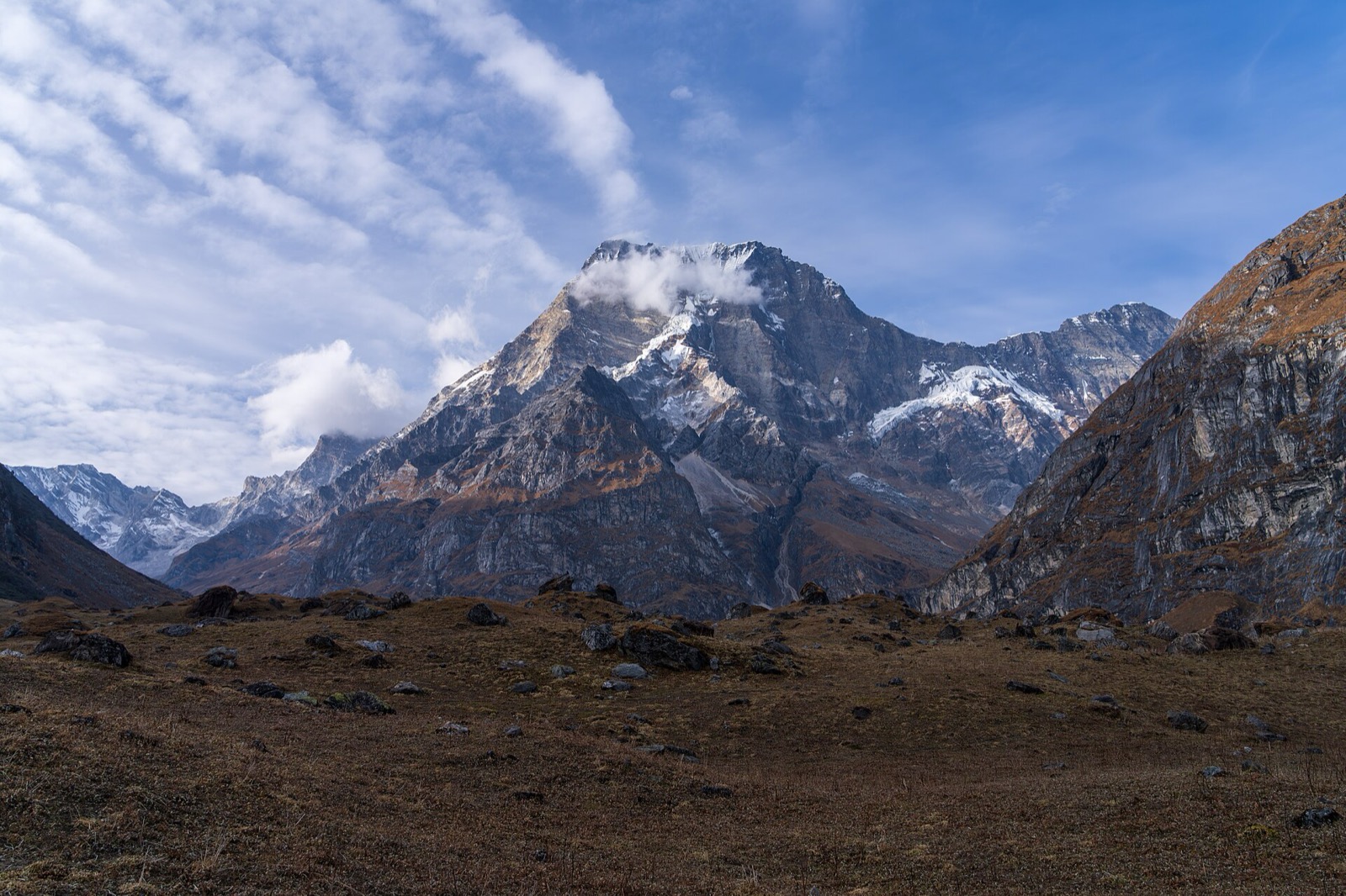 A snow-dusted rocky peak with cloud on its summit rises above a brown alpine meadow