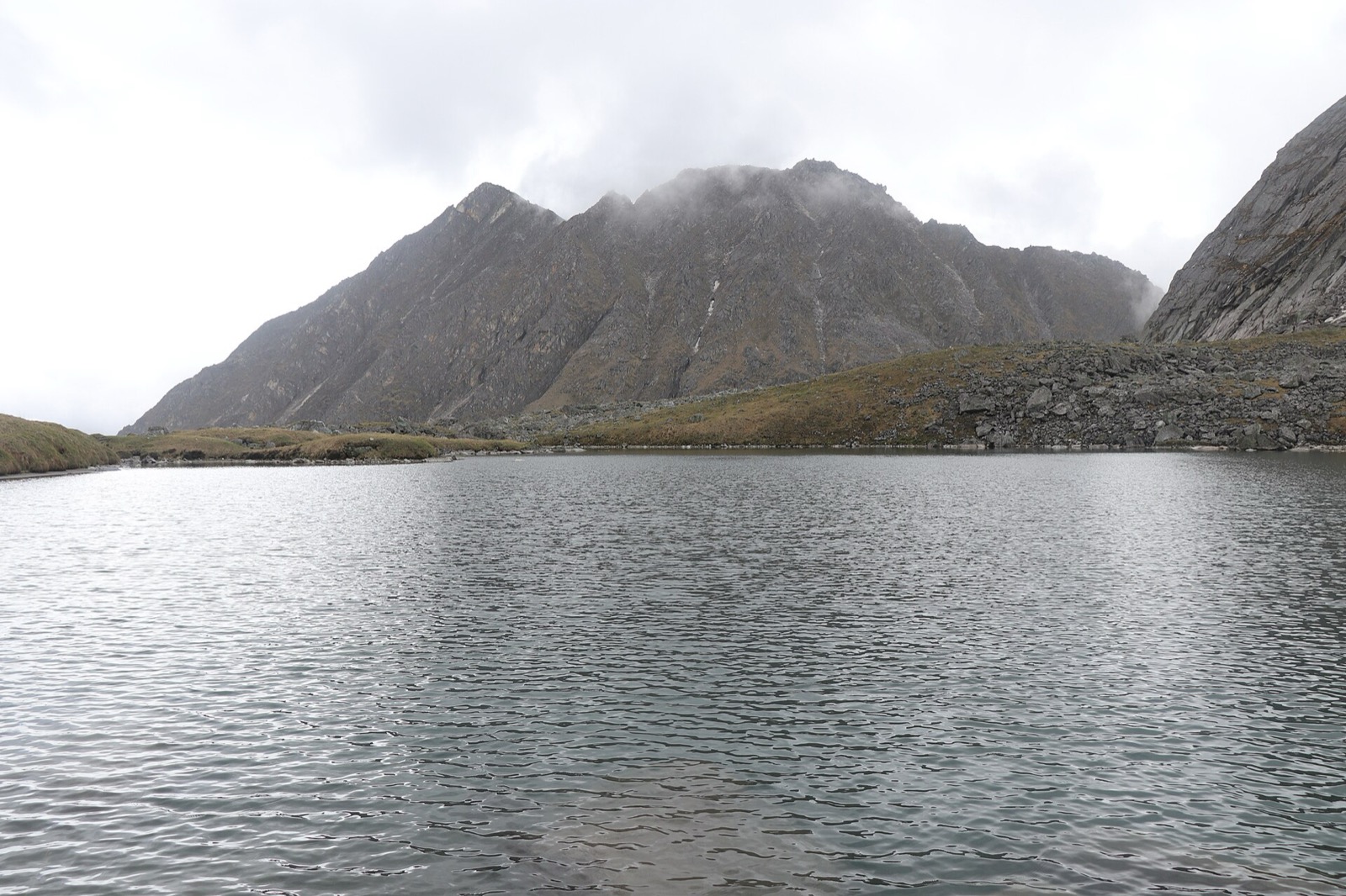 A still grey lake lies below a bare, cloud-capped ridge of dark rock