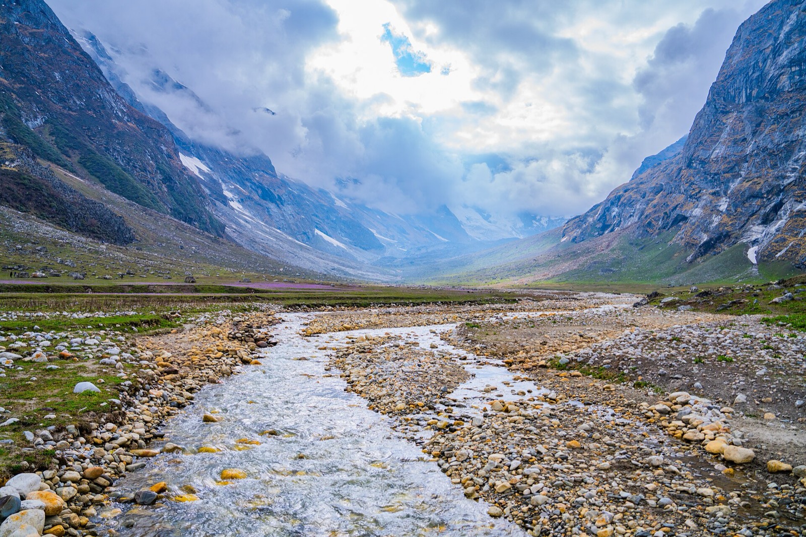 A braided glacial river runs down a wide valley between steep rocky mountains under cloud, with snow patches on the slopes